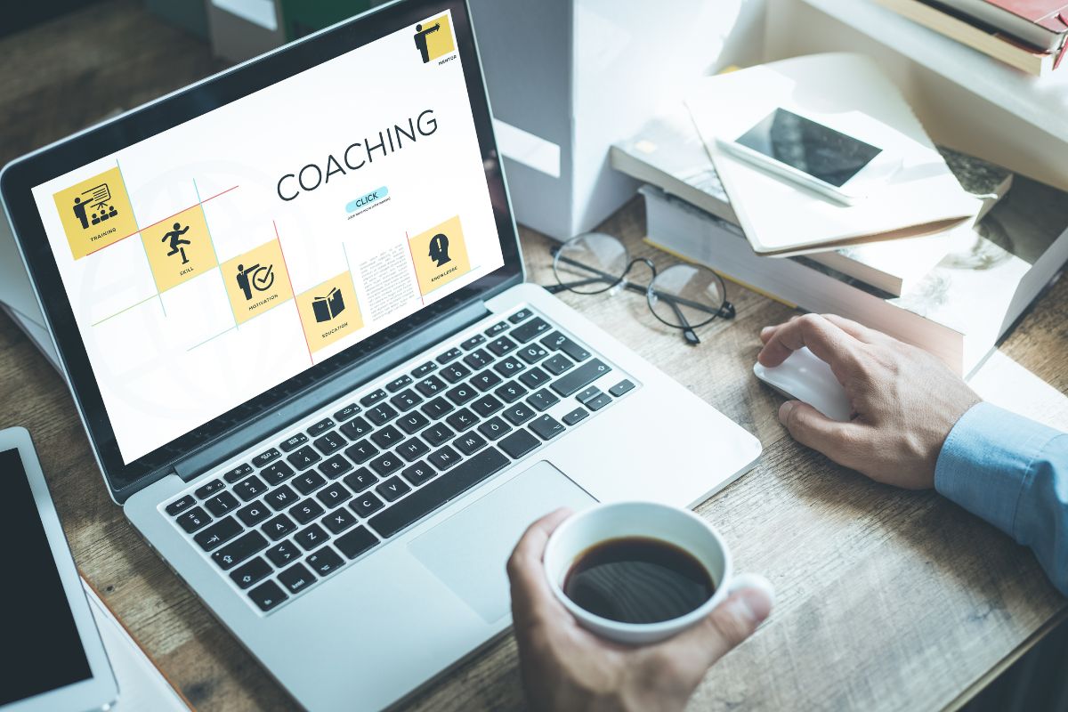 The image shows books, a phone, an eyeglass, and a man's hand using a laptop while having a coffee indicating that offering high-ticket coaching can be a very profitable side business.