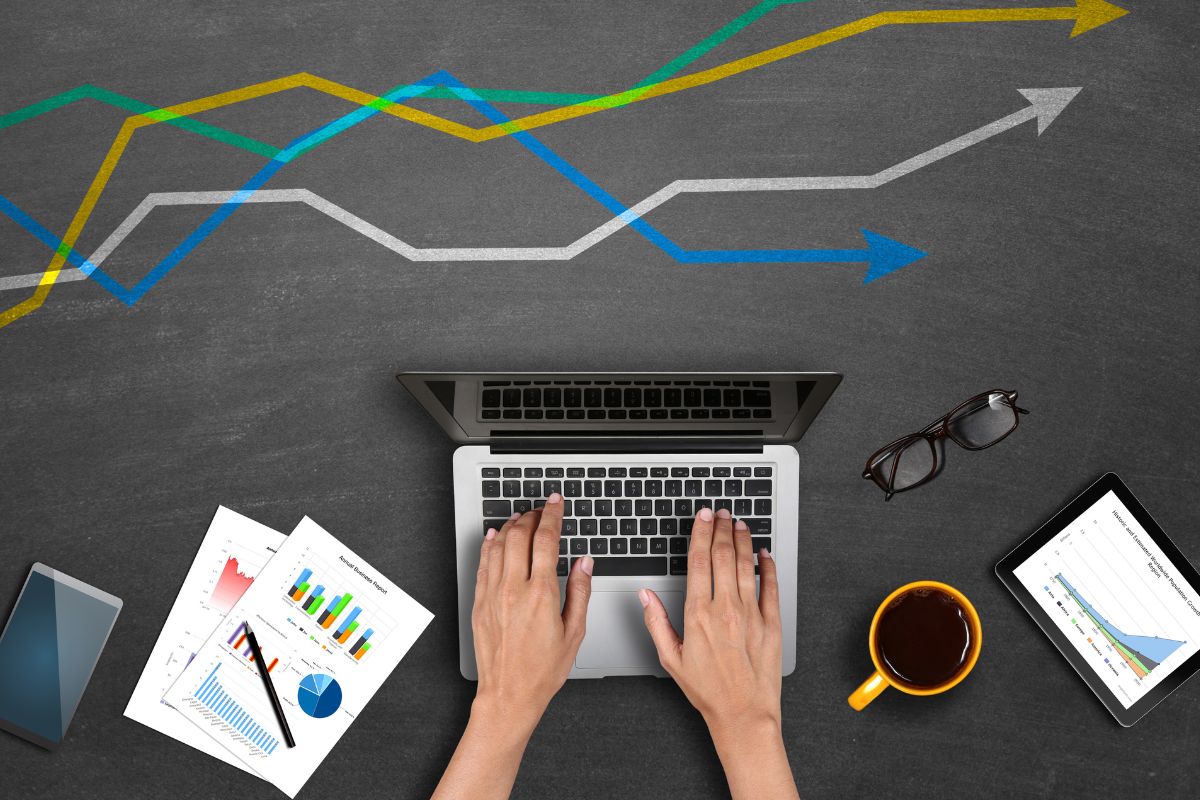 The photo tells about papers, a phone, a tablet device, an eyeglass, and a girl's hand using a laptop which means selling exclusive research reports in trending industries that help them save time and can be a way to make money online.