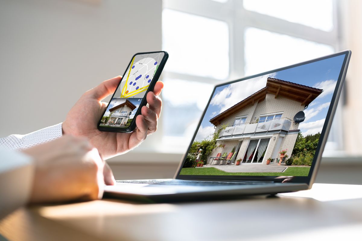 The picture tells about a laptop with a house image on the screen and a man's hand holding a phone with GPS which means buying and flipping digital real estate can be a way to make extra money from home.