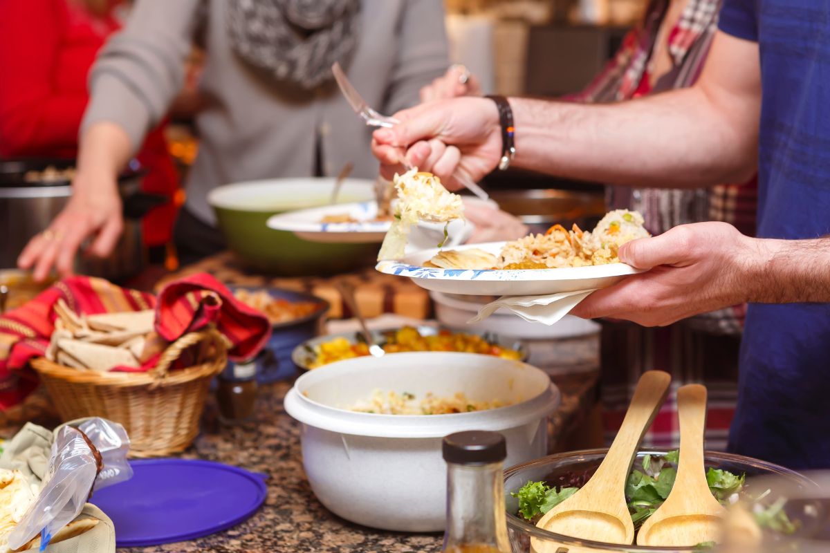 The picture shows food on the tables and a group of people getting onto their plates which means to host potlucks is an easy way to keep your social life active without draining your budget.