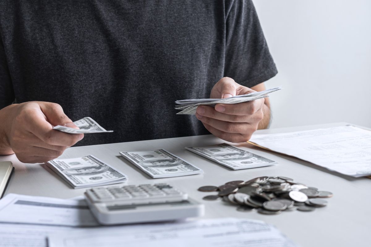 The photo shows coins, a calculator, papers, and a man dividing the dollar bills into groups which means using the rich first strategy to make financial moves early.