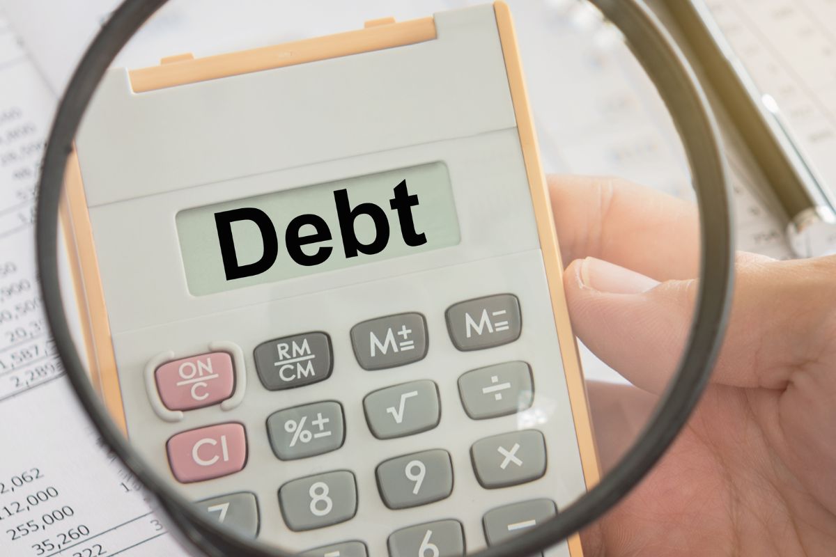 The picture shows papers with numbers, a pen, and a man's hand holding a magnifying glass and a calculator referring to the use of velocity banking to pay off high-interest debt means more money for investing and building wealth.