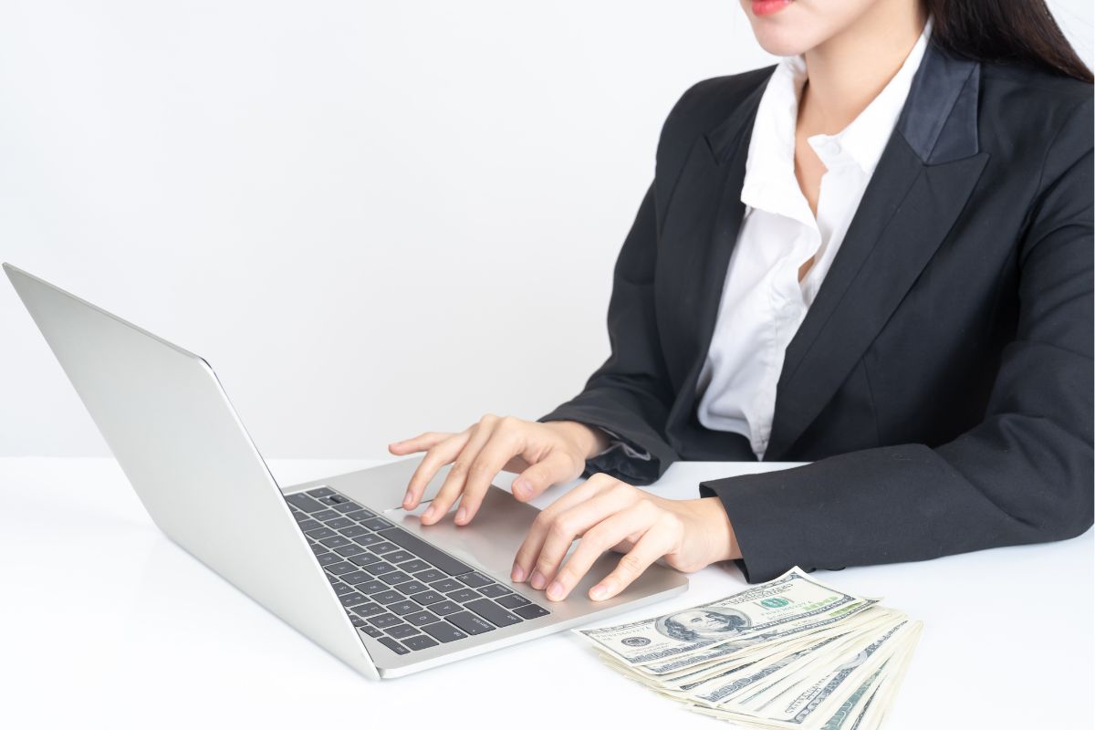 The photo shows dollar bills on the table and a girl using a laptop referring to the use of the invisible money trick to ensure steady financial growth and easier to build wealth.
