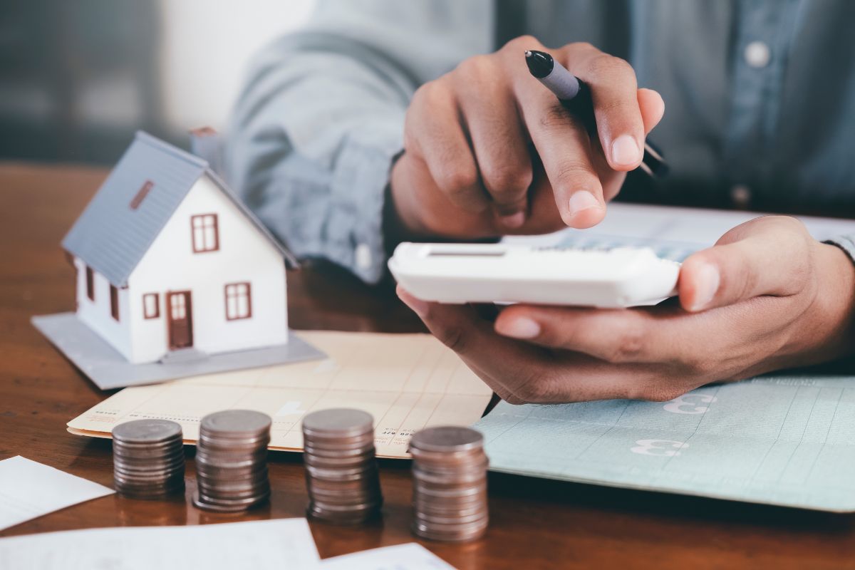 The image shows coin stocks, a bank book, a mini house, and a man's hand calculating which means avoiding penny-wise, dollar-foolish habits, and saving money in the right places has a much bigger impact than skipping a daily treat.