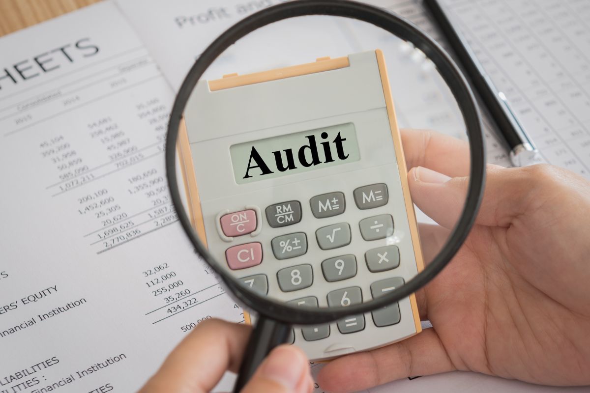 The photo tells about papers with notes, an open, and a man's hand holding a magnifying glass and a calculator which means to audit spending every 90 days to help eliminate waste and keep money working in the right places.
