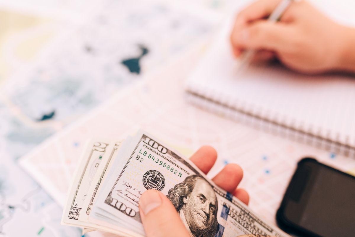 The image shows a phone and a man's hand holding dollar bills while writing in the notebook which means that using future budgeting impacts your long-term wealth.