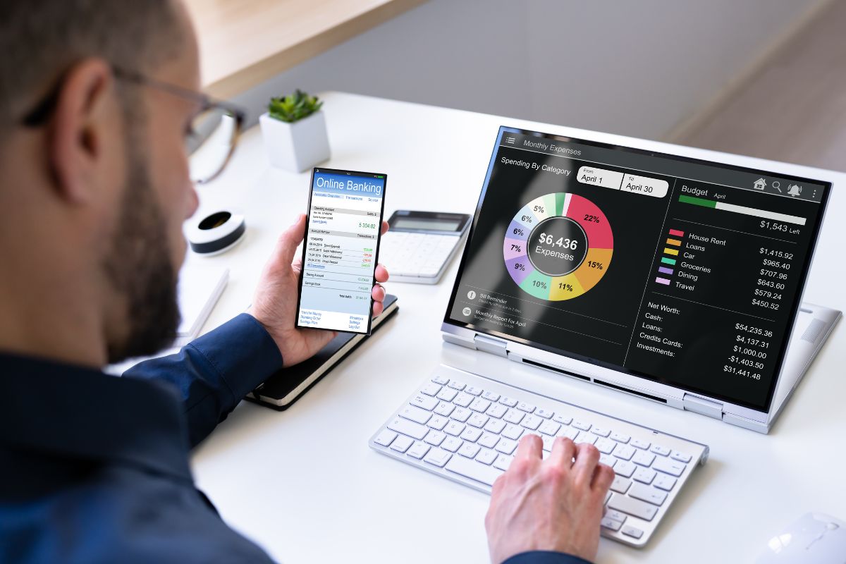 The photo tells about a man using a laptop while holding a phone which means using a budgeting app makes it easy to see where your money goes.