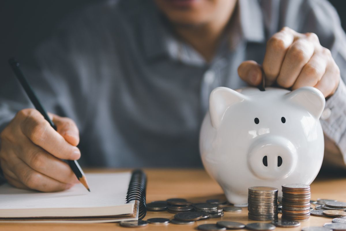 The picture talks about coin stocks and a man's hand and writing on the notebook while dropping a coin into a piggy bank indicating that turning savings into a game keeps you motivated and turns saving into something to look forward to.
