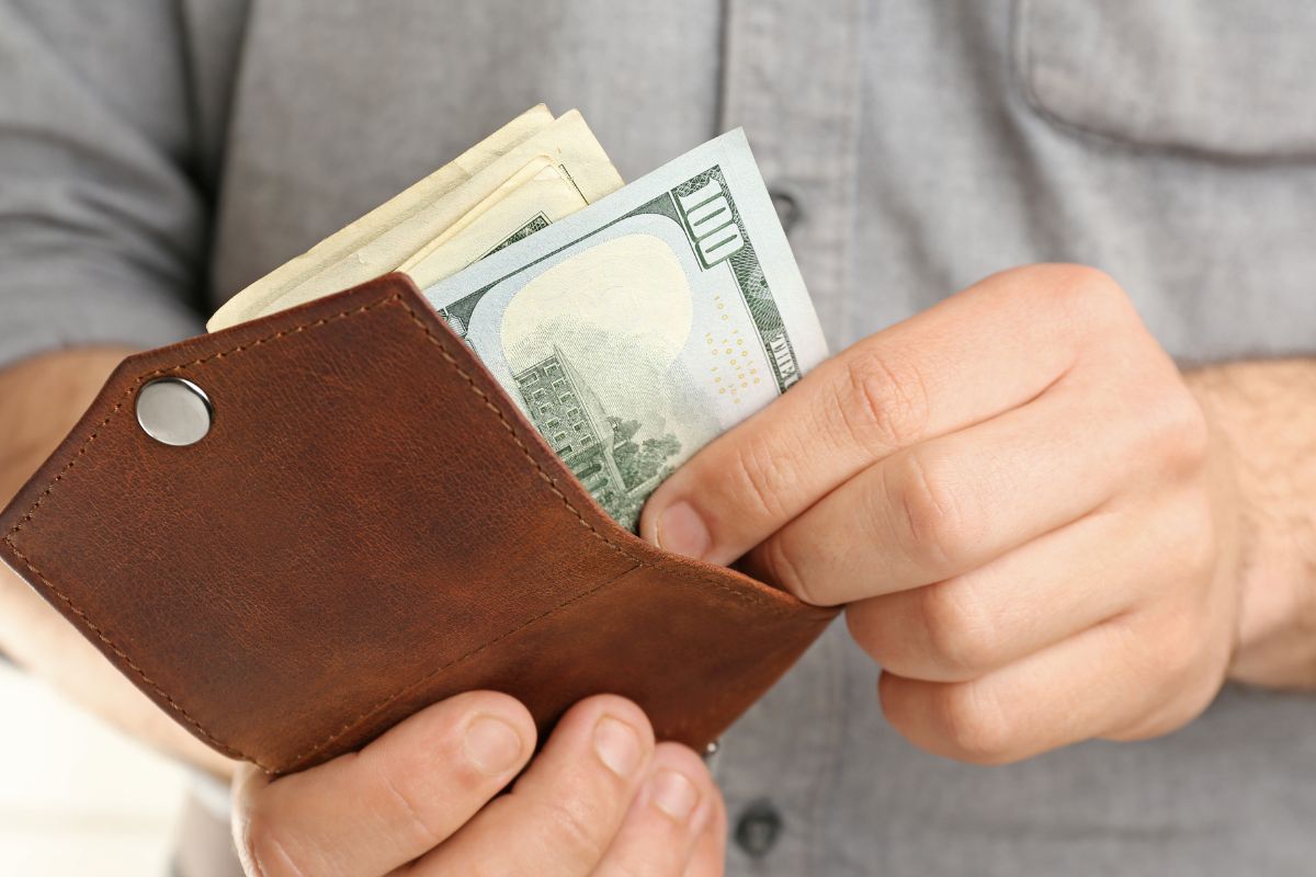 The image shows a man's hand putting back the dollar bills inside his wallet which means creating a no-spend day helps you reset your spending habits and see where your money normally goes.