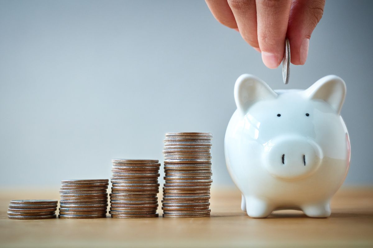 The picture shows stock coins and a man's hand dropping coins inside a piggy bank which means to gamify savings with a friend, it is easier to stick to money goals when you have someone cheering on you.