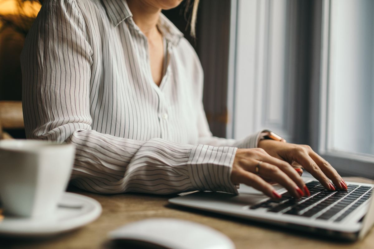The photo shows a coffee, a mouse, and a girl using a laptop referring to becoming a virtual assistant as a good way to work from home while learning useful skills. 