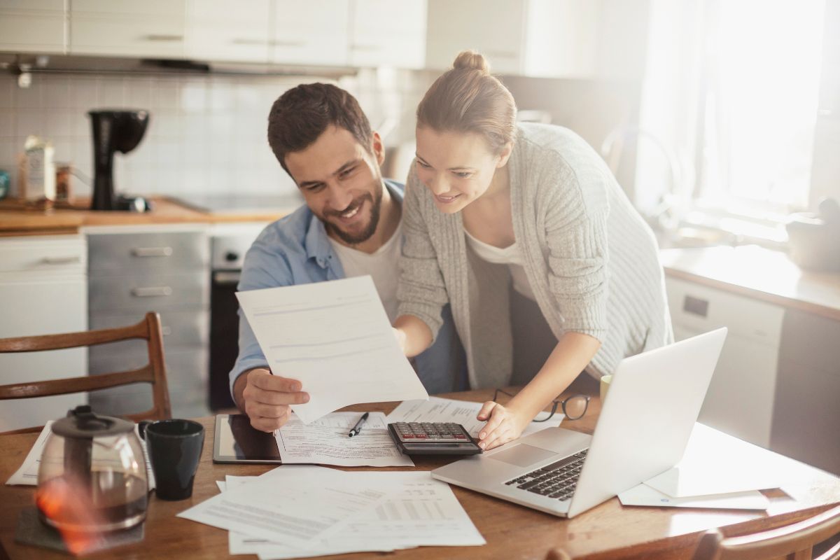The photo shows papers on the table, a laptop, a coffee, a mug, a calculator, and a couple of checking papers referring to holding yourself accountable makes it easier to stick to your budget.