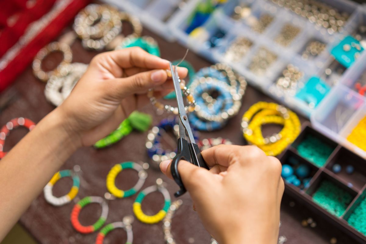 The image tells of beads in the container and a girl's hand cutting the wire of the finished product bracelet which means selling handmade products on online platforms can control pricing and production while reaching a large audience.
