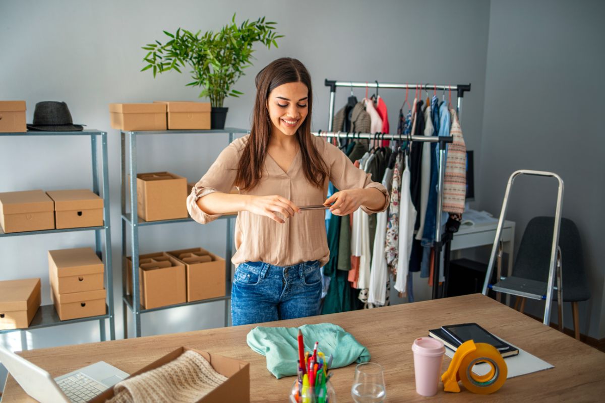 The image tells about boxes on a rack, hung clothes a laptop, a notebook, and a girl taking a picture of clothes which means flipping items from thrift stores on online platforms is a simple way to make money and to sell online.