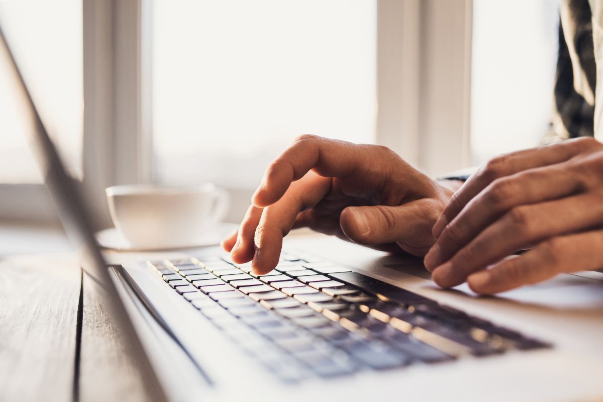 The picture shows a cup of coffee and a man's hand using a laptop which means publishing low-content books on Amazon KDP is a great passive income idea because once the book is listed, it can sell for a year.