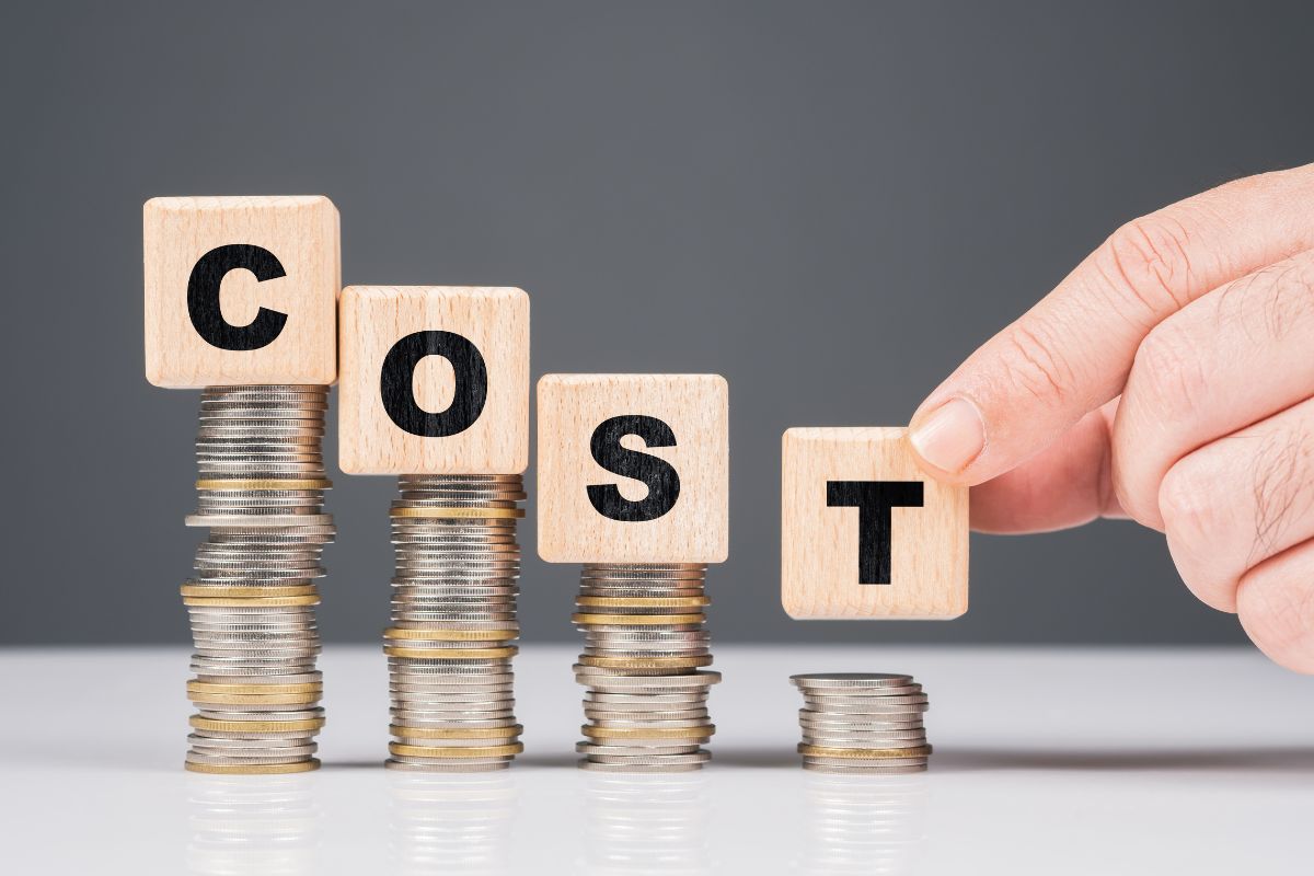 The picture tells about stock coins, wooden cubes with letters, and a man's hand holding the last letter referring to reduced fixed costs frees up money for savings and important expenses.