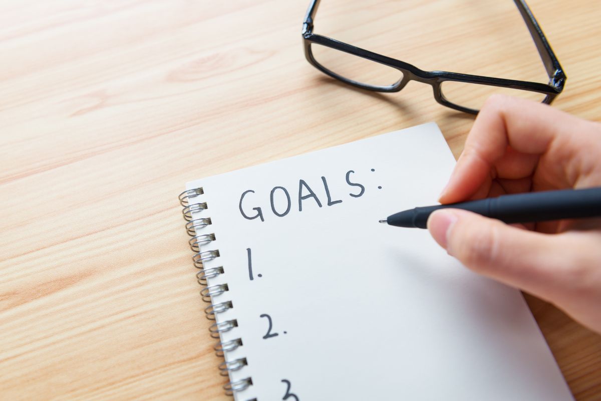The photo shows an eyeglass and a girl's hand and writing on the notebook indicating that setting clear financial goals keeps you moving in the right direction.