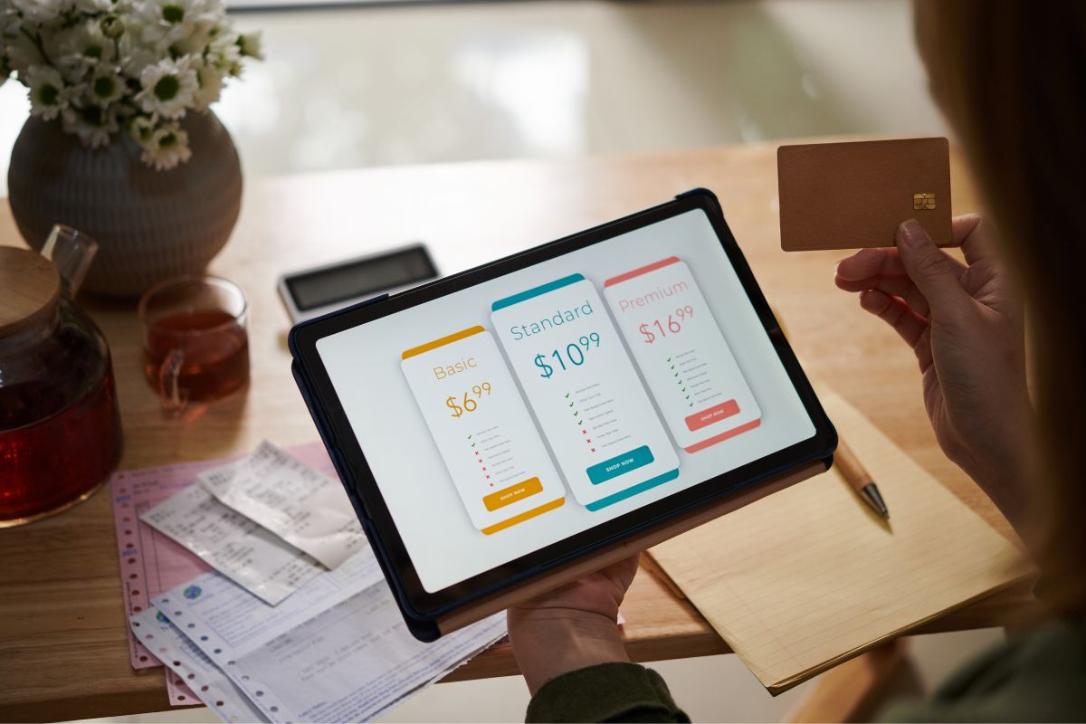 The image tells about a flower vase, tea, receipts on the table, and a girl holding a tablet device with a screen display and a credit card which means discretionary spending can add up if not watched, budgeting for fun lets you enjoy life without guilt.