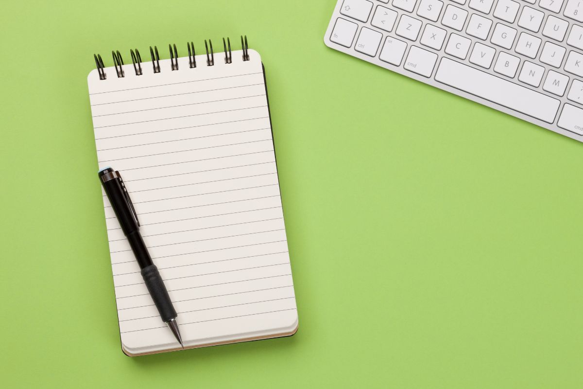The image shows a keyboard, a pen, and a notebook which means to create a spending temptation list helps break impulse buying habits and stick to the challenge.