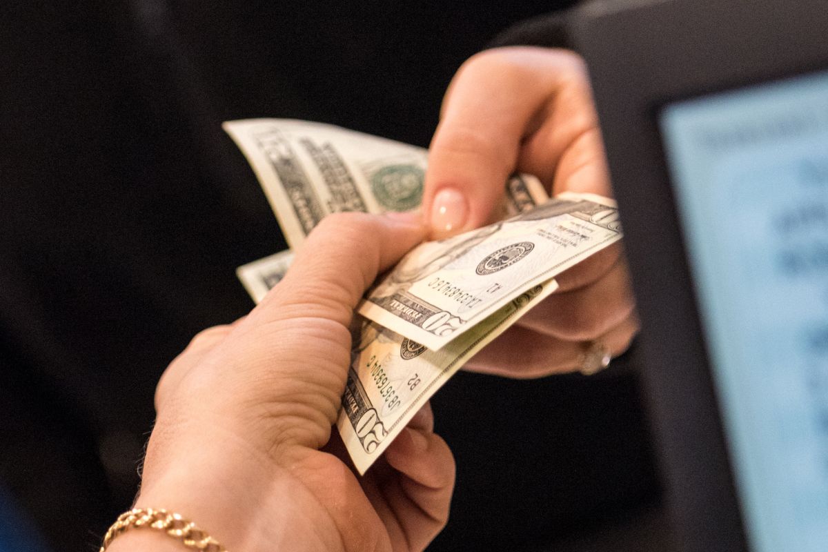 The photo tells about a customer's hand handing the dollar bills to the cashier's hand which refers to paying for needs in cash and wants to prevent overspending and keeps you in control of your money.