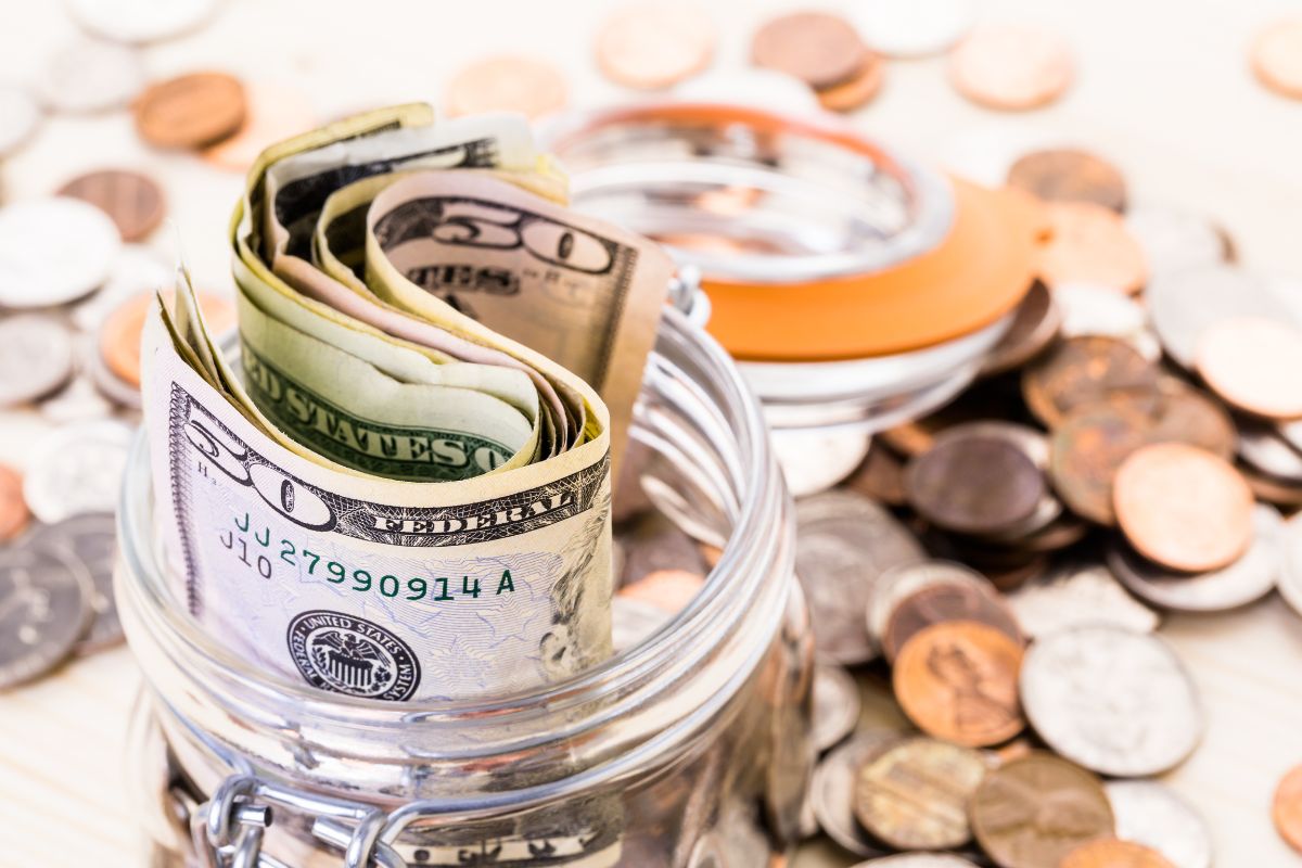 The image shows coins and a jar filled with coins and dollar bills which means setting up a fake bill to pay yourself first ensures you put money toward goals instead of wasting it.
