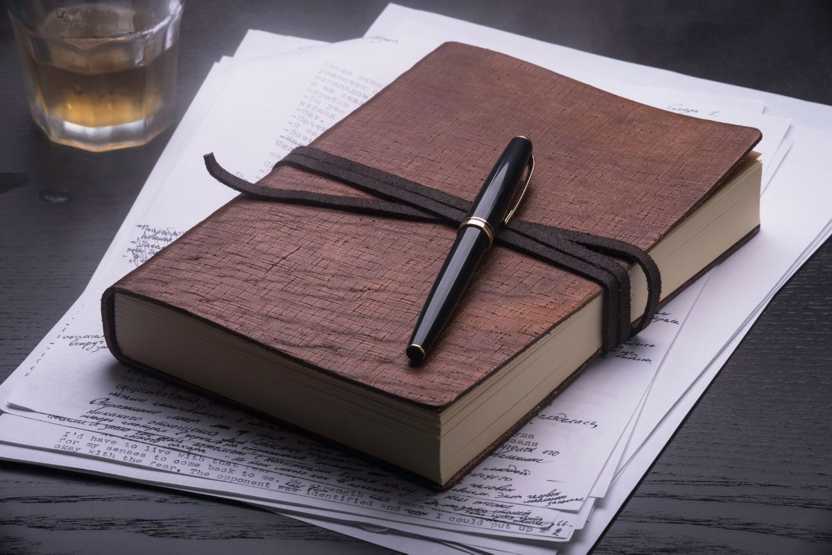 The photo shows a glass of drink, papers, a pen, and a notebook indicating that keeping a success journal of money saved helps you track progress and stay focused.