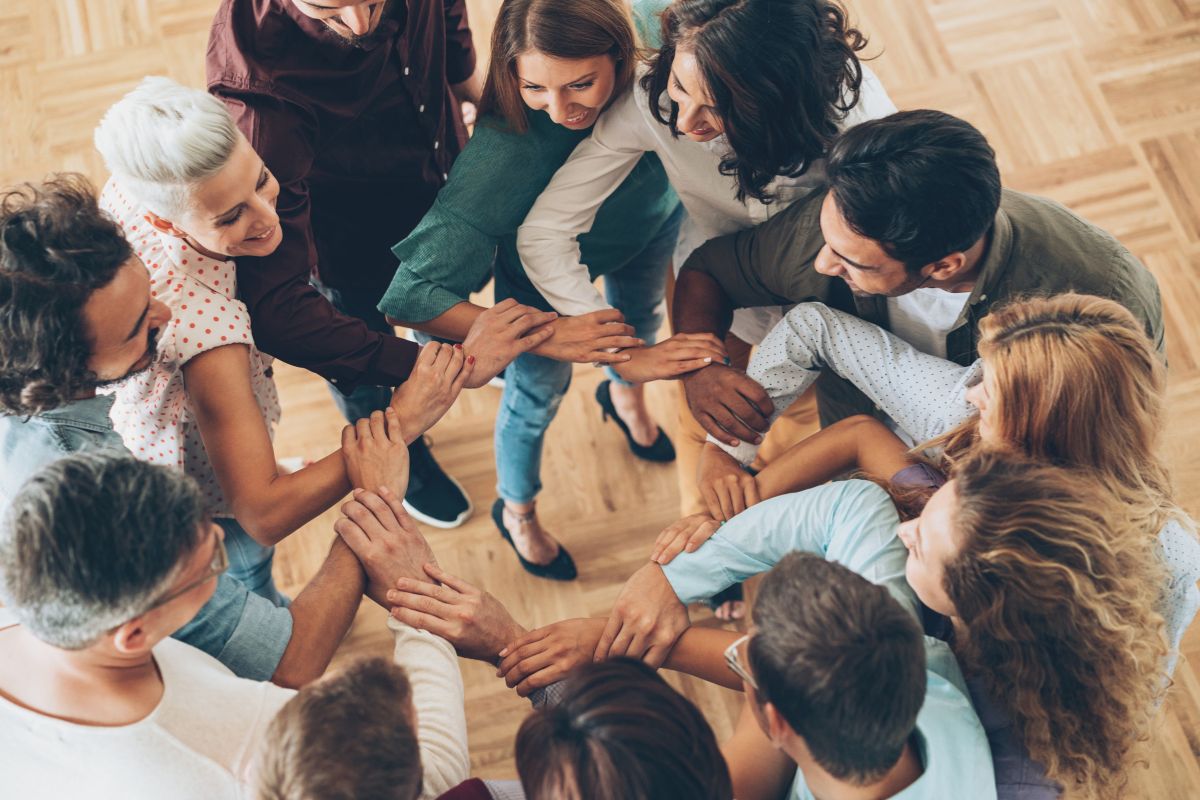 The photo shows a group of people joining hands together while forming a circle referring to how surrounding yourself with like-minded people helps you stay committed to smart money habits.