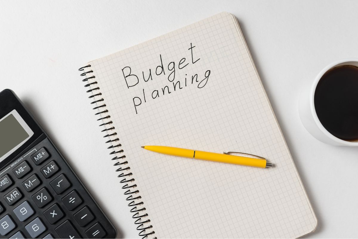 The photo shows a coffee, a calculator, a pen, and a notebook with notes which means creating and sticking to a budget helps you avoid living paycheck to paycheck.