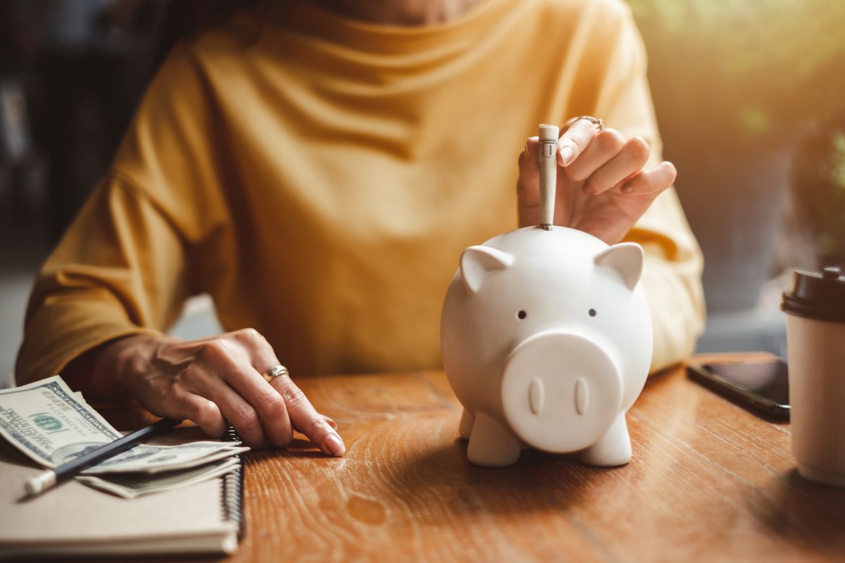 The picture shows a coffee, a notebook, a pen, dollar bills, and a girl dropping a dollar bill in a piggy bank referring to paying yourself first makes reaching financial independence much easier.