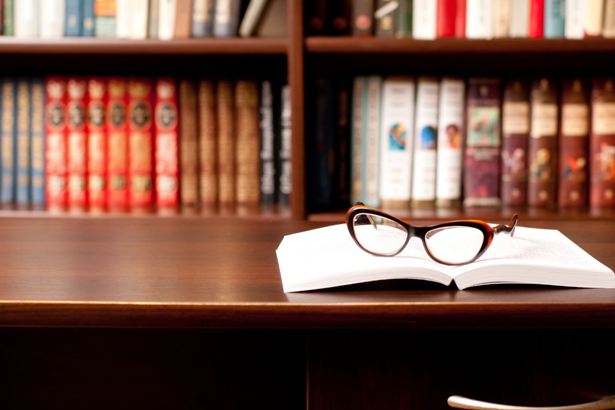 The photo tells about books on the shelves, a book on the table, and an eyeglass which means using the library for books helps cut unnecessary spending while still enjoying quality entertainment.
