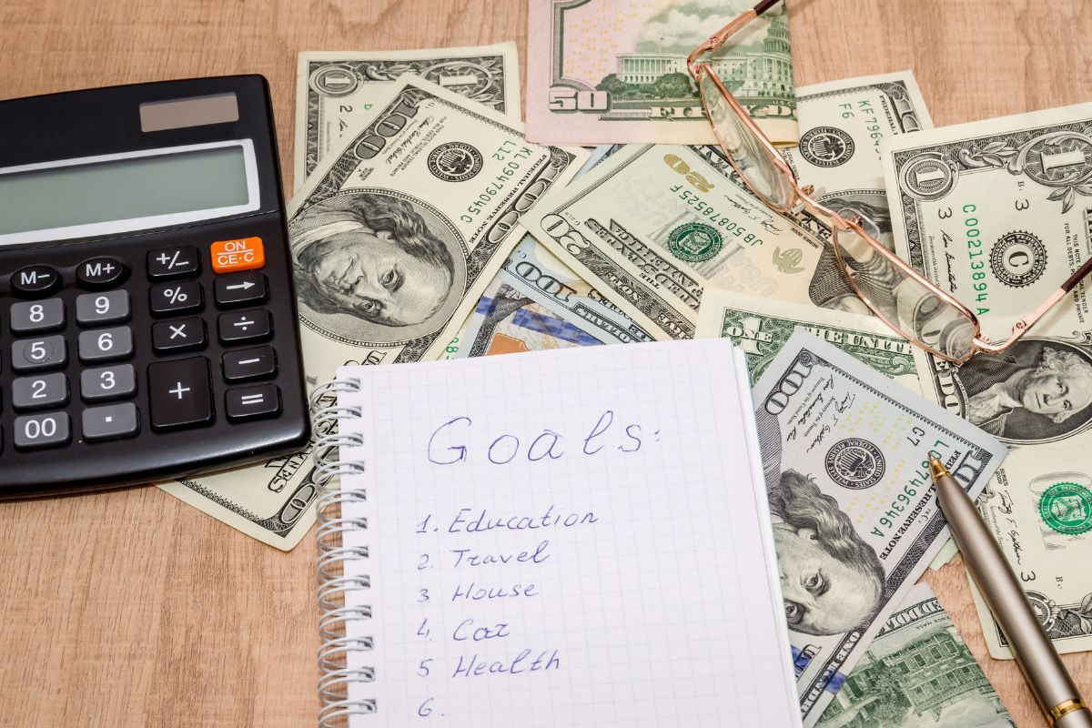 The picture tells about dollar bills, a calculator, an eyeglass, a pen, and a notebook with notes referring to setting clear financial goals helps adjust plans and stay motivated.