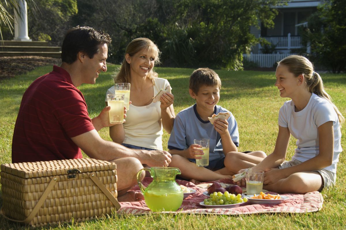 The image shows a group of families enjoying their food at the park referring to having a picnic at a local park as a great way to take a break from eating out and save some money.