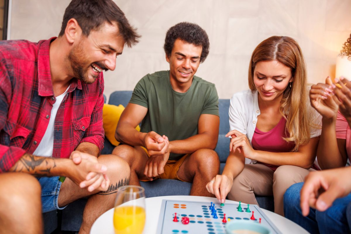 The picture shows a group of people playing a chess game with juice on the table indicating that hosting a game night with board games helps avoid the cost of going out.