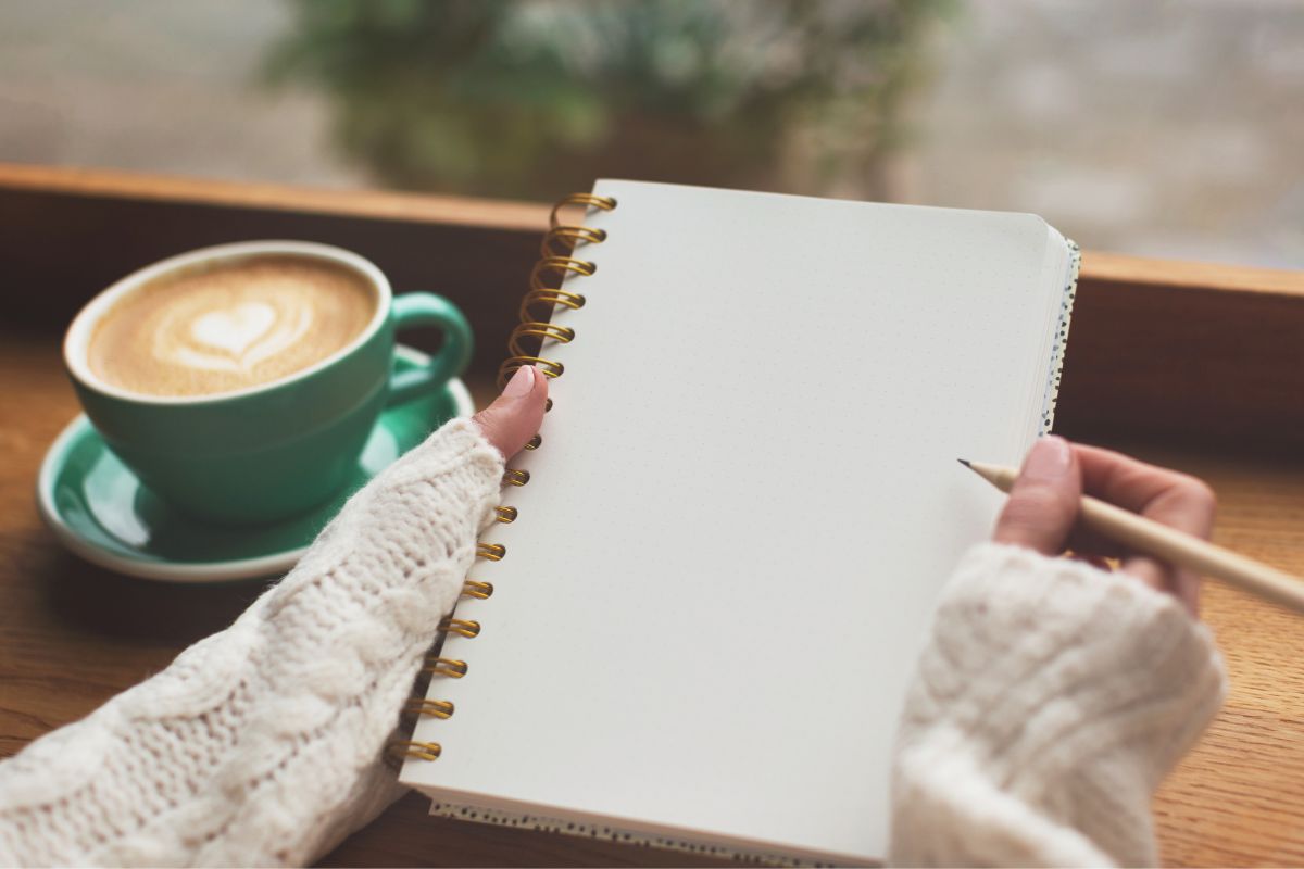 The image shows a coffee and a girl's hand holding a notebook and a pen referring to writing in a journal helps clear your mind and might even turn into a new hobby.