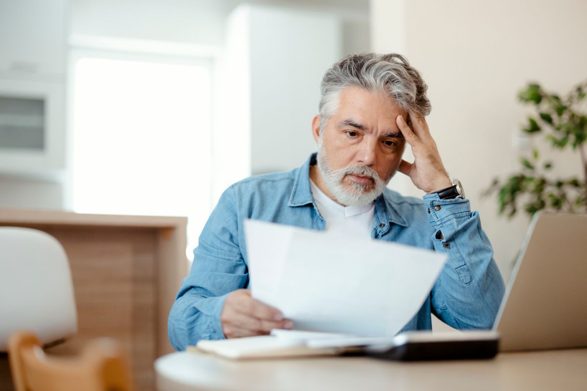 The photo is about a laptop, a notebook, a calculator, and a man holding a piece of paper while holding his head which means that solving high-value problems brings financial success and avoids stress.