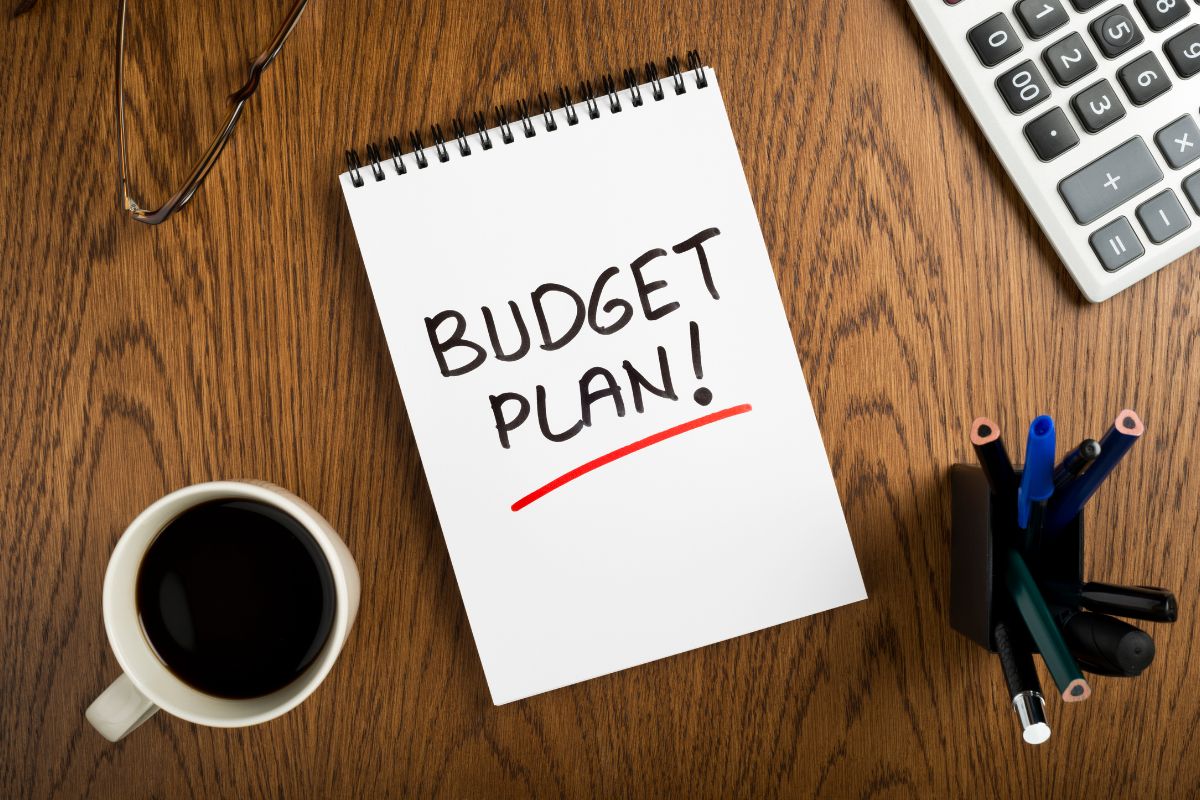 The picture tells about a coffee, a calculator, pens, and a notebook with notes which means to plan your upcoming budget helps you stay on track with your money goals.