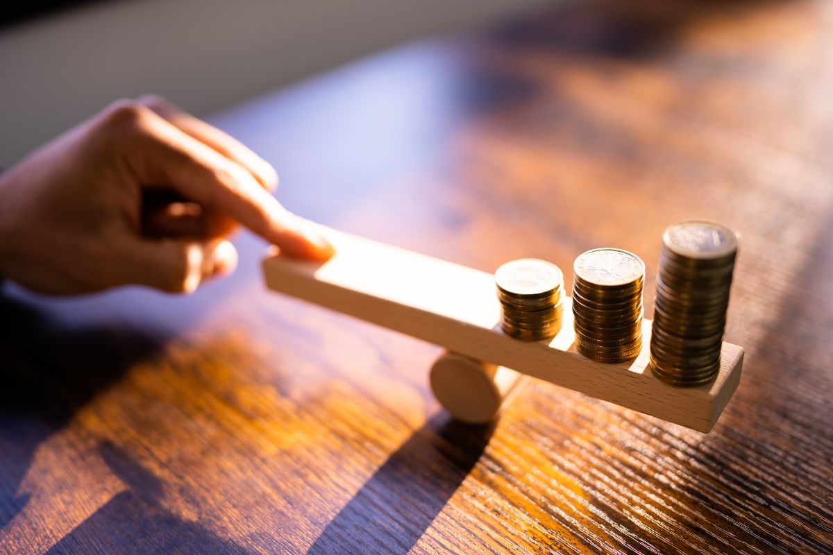 The image shows a weighing wood with coins and a man's hand on the other side means leveraging other people's money to grow wealth faster which leads to bigger results.