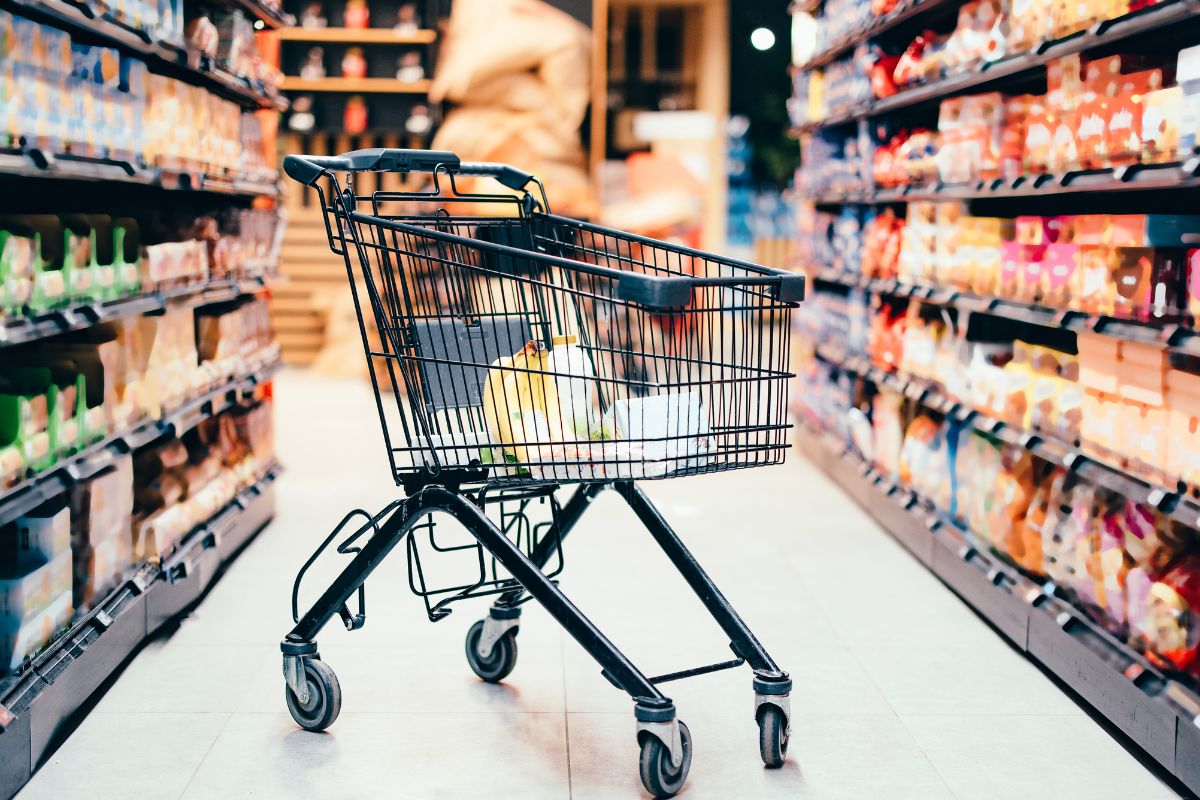The picture of a product display and a cart with items inside which means to limit grocery shopping to only essential keeps grocery costs low while still allowing for balanced meals.
