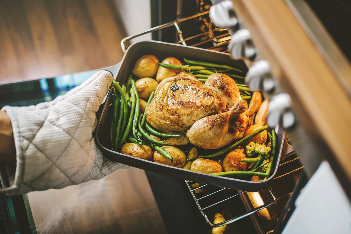 The picture tells about a girl's hand putting a tray of chicken with vegetables in the oven which means to batch cook and freeze meals saving time and preventing waste while also making busy nights easier.