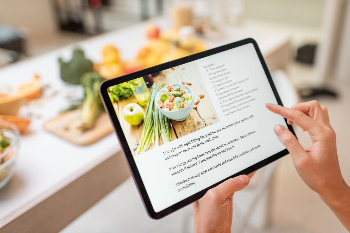 The photo shows ingredients behind and a girl's hand holding a tablet device with a screen display which means using online recipe generators helps avoid waste and gives new ideas for using pantry staples.