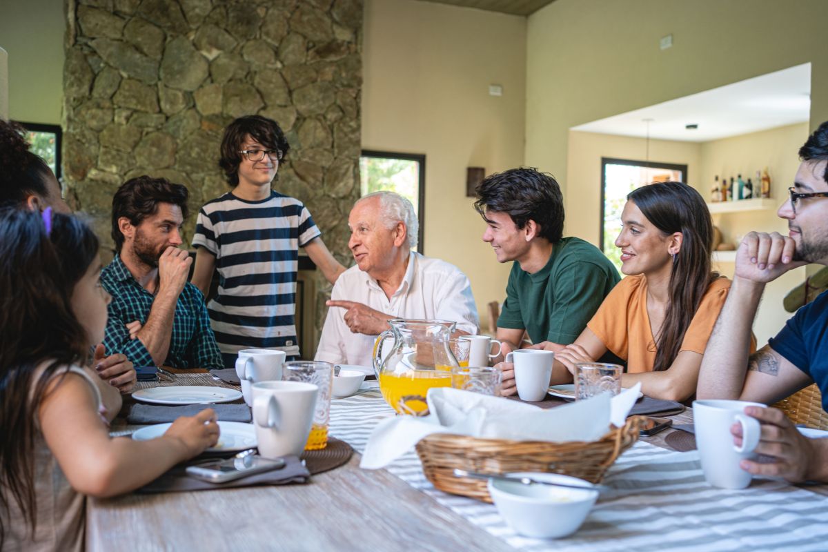 The picture of mugs, plates, juice, a basket on the table, and a group of family talking together which means getting the whole family involved in meal planning teaches good habits while keeping grocery costs low.
