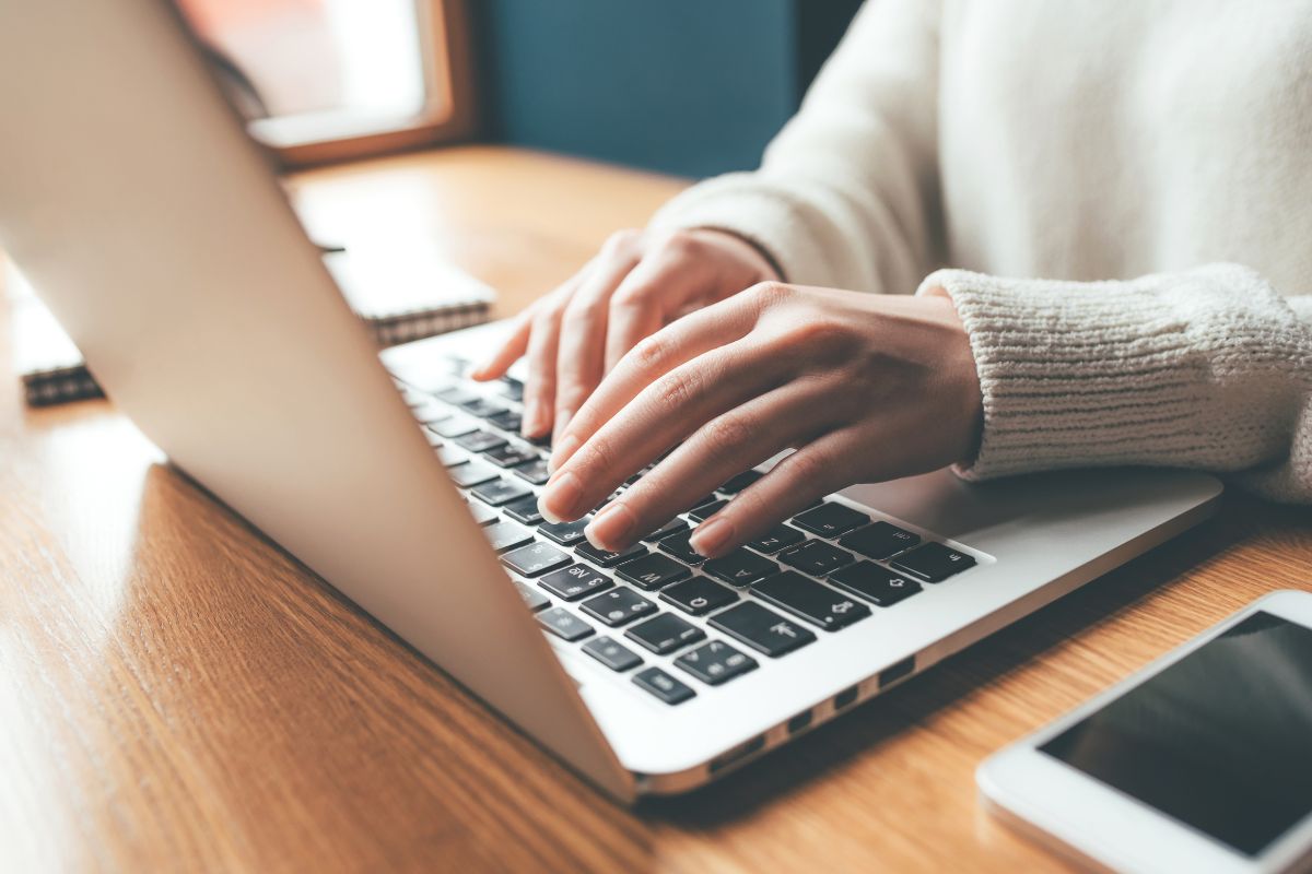The photo tells about a phone, a notebook, and a girl's hand typing on the laptop referring to the focus on creating multiple streams of income for more chances to grow wealth and stay financially strong.