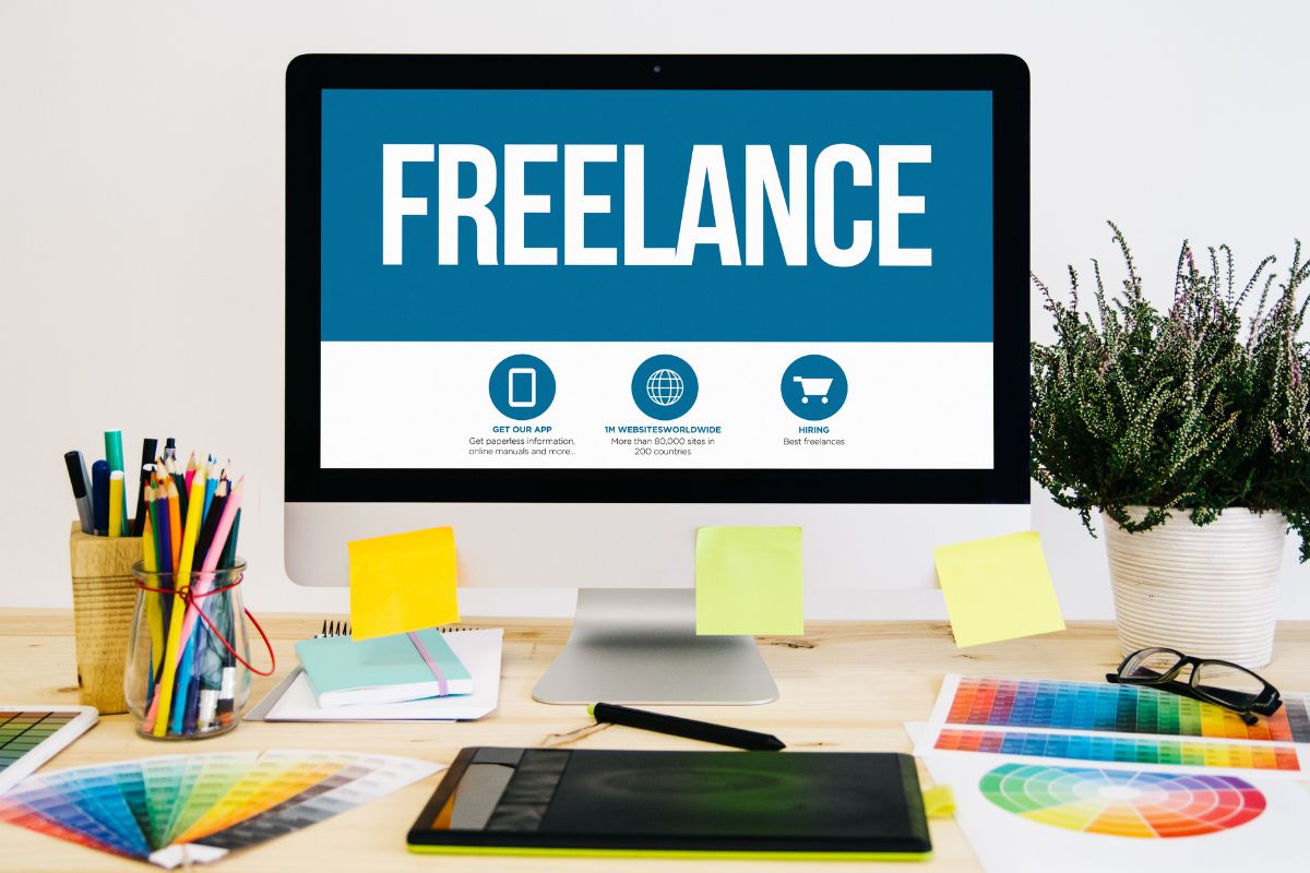 The photo shows pens, colors, papers, notebooks, a plant, and a monitor with a screen display indicating that opening doors to freelance give you more financial security and freedom.