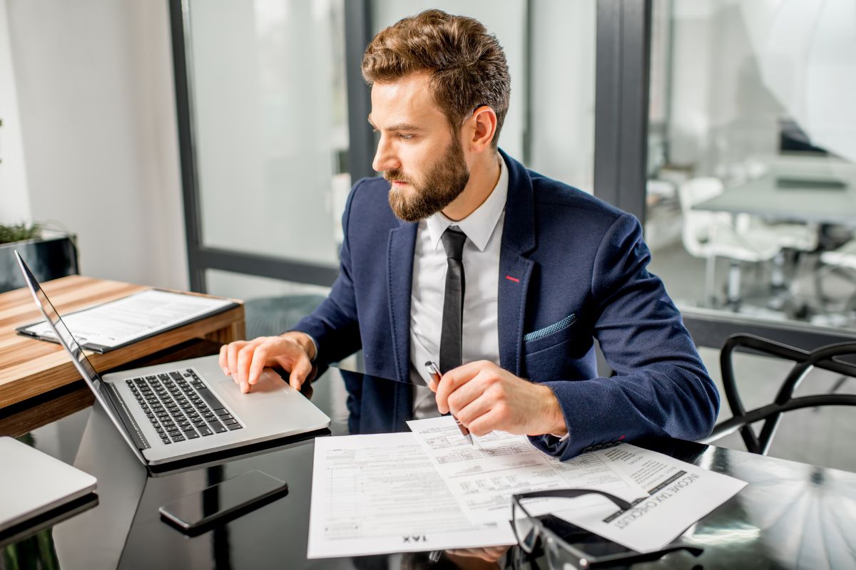 The photo shows papers on the table, an eyeglass, a phone, and a man in a suit using a laptop to learning new skills boosts productivity and efficiency makes you stand out, and increases your earning potential.