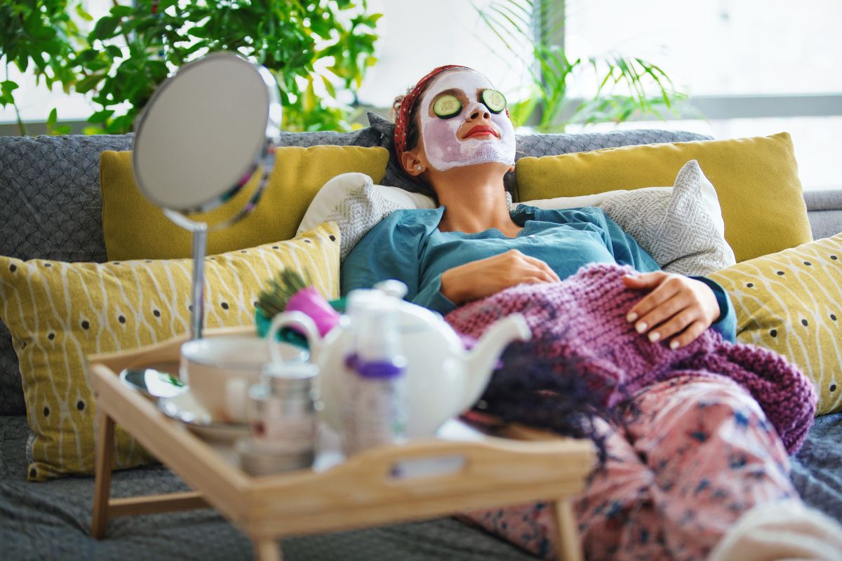 The image of a mirror, tea cup, and a girl with a face mask and cucumber slice on her face lying on the couch which means having a no-spend weekend can help reset your habits and keep more money in your pocket.