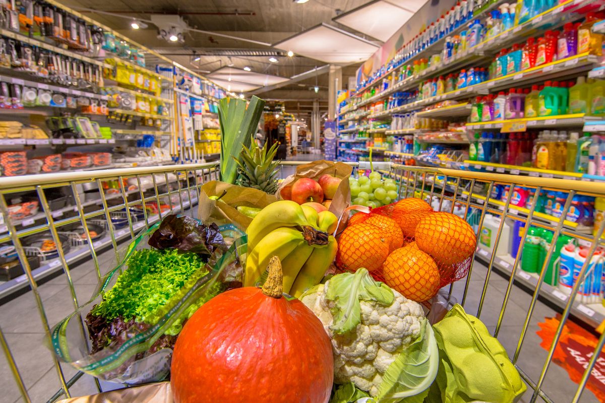The image shows a product display and a cart filled with vegetables and fruits referring to limiting grocery store trips that can help make groceries last longer.