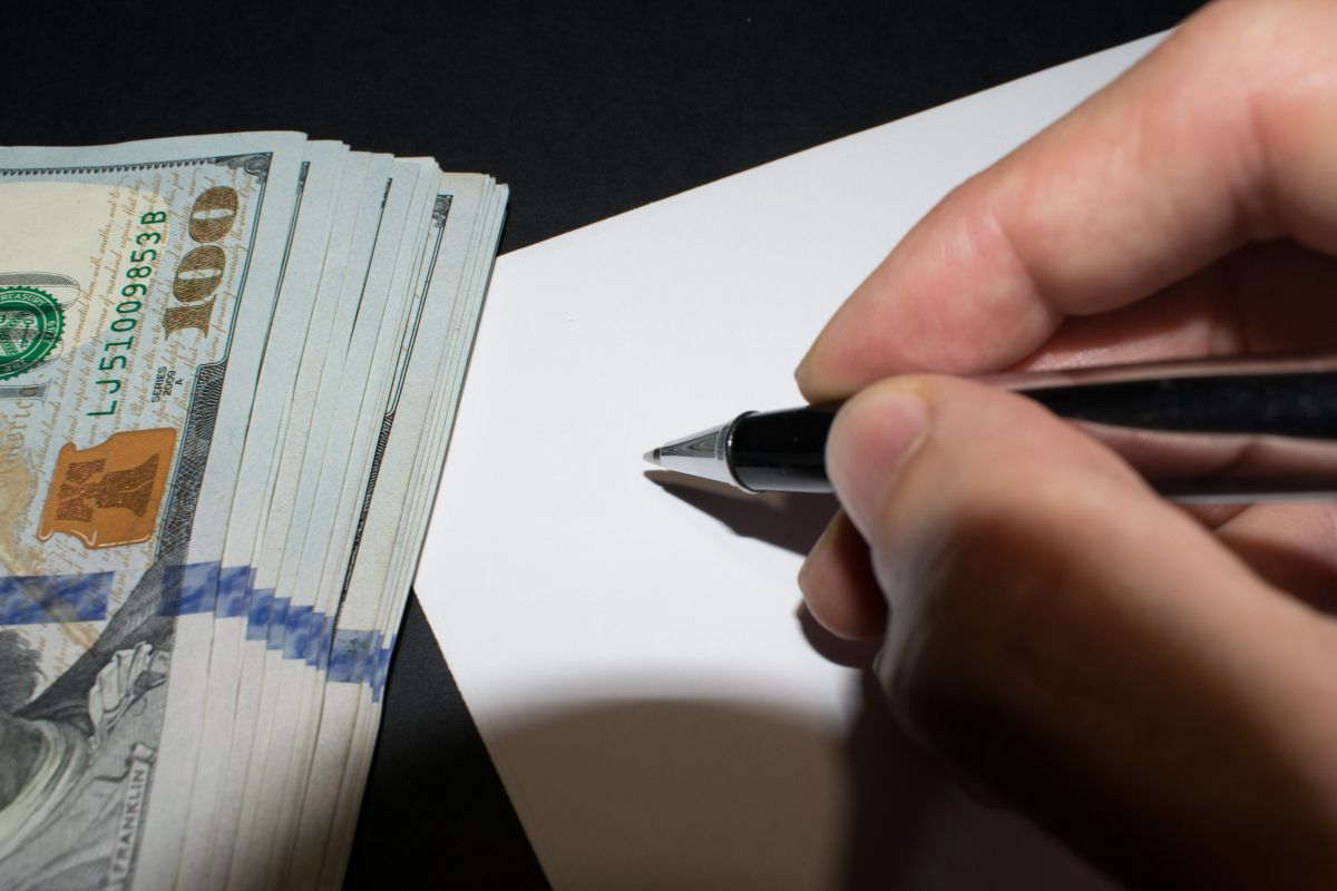 The image shows dollar bills and a man's hand and writing on the piece of paper referring to schedule money check-in days keeps your finances in good shape.