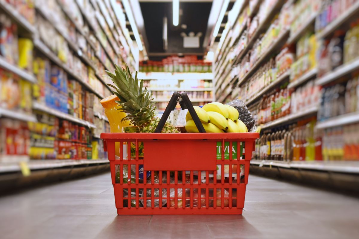 The photo shows product displays and a basket with fruits, vegetables, and more which means that delaying grocery shopping by a few days saves money over time.
