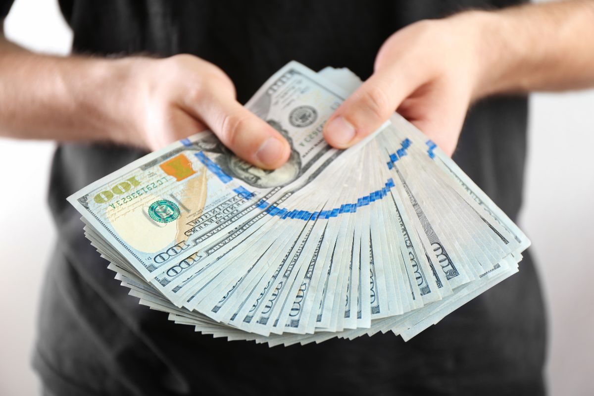 The image shows a man's hand holding dollar bills referring to challenging yourself to save your first $100k to help you reach your financial goals faster.