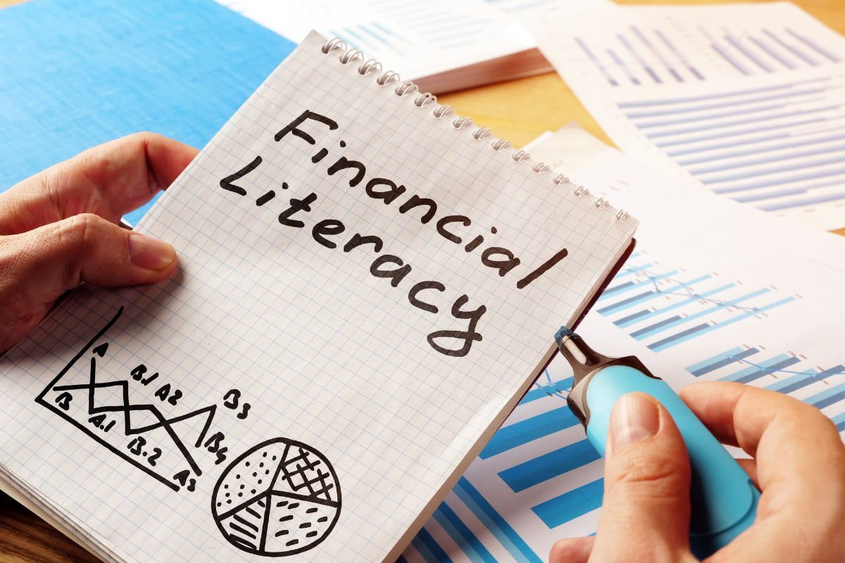 The photo shows papers with graphs and a man's hand holding a notebook with notes and a marker indicating that learning new skills improves financial literacy and can increase your wealth over time.